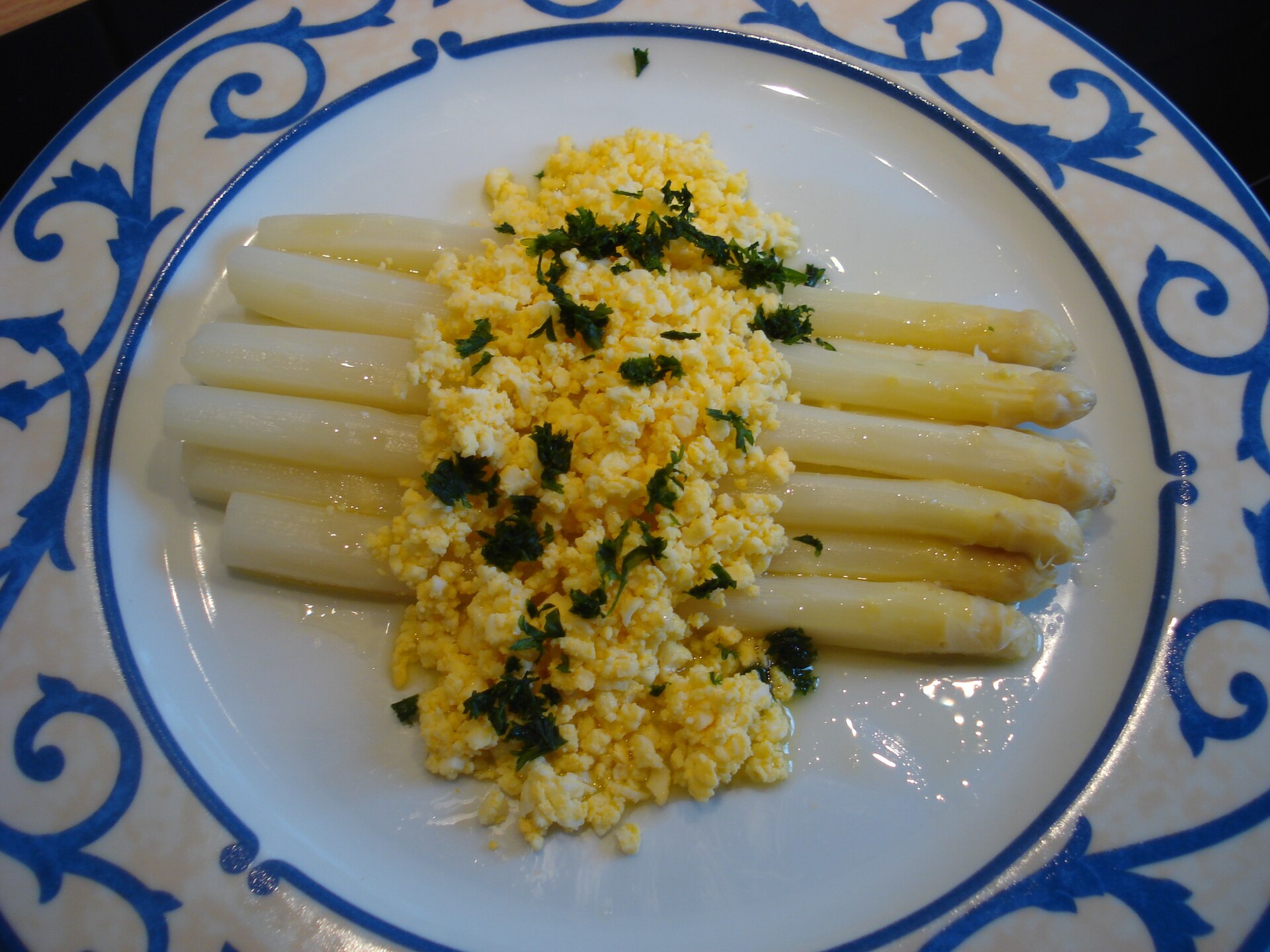 White asparagus spears on a blue-patterned plate, covered with chopped hard-boiled egg, melted butter and parsley in the Flemish style