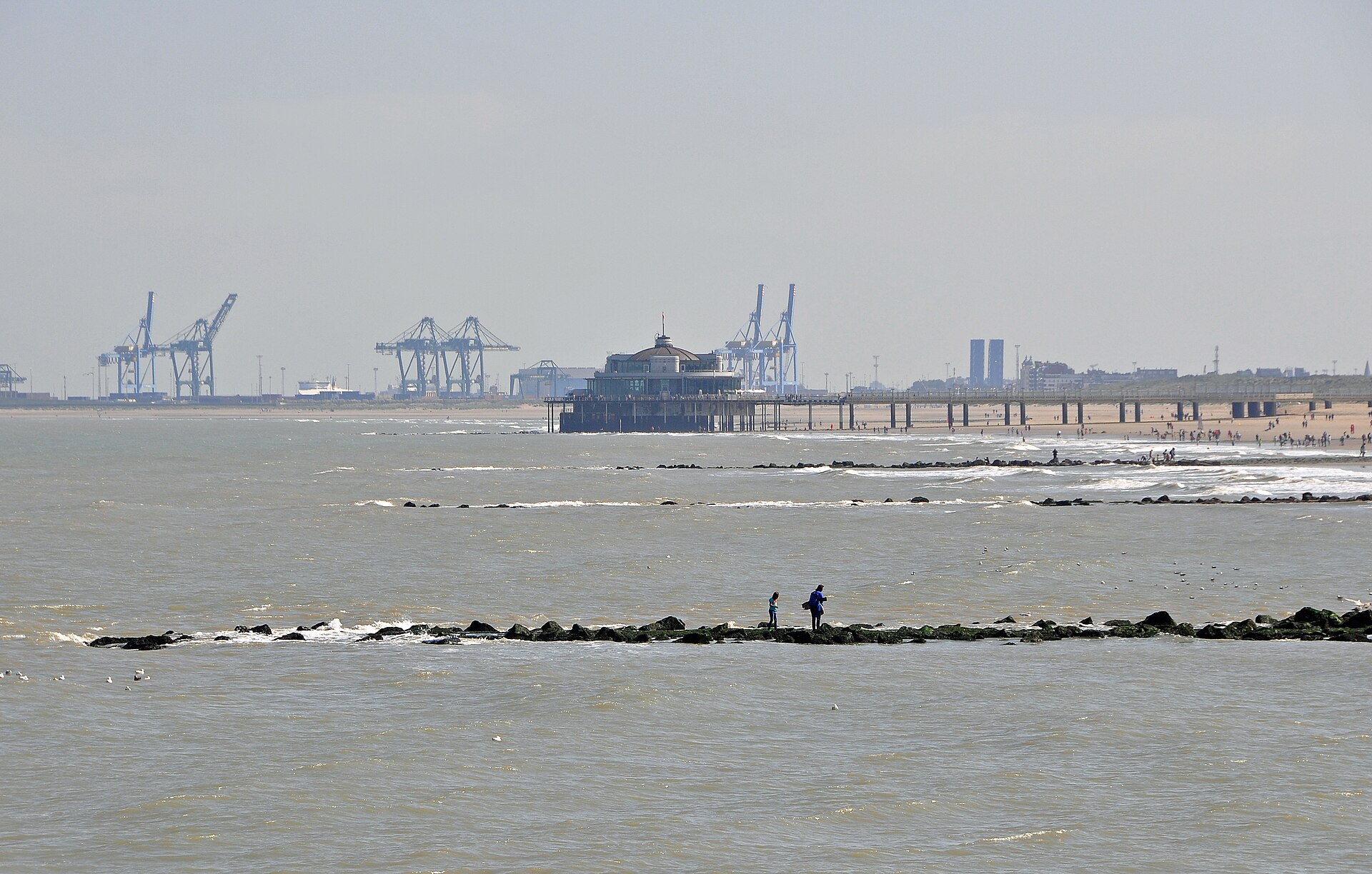 The pier at Blankenberge seen from the beach with the container cranes of Zeebrugge port behind it