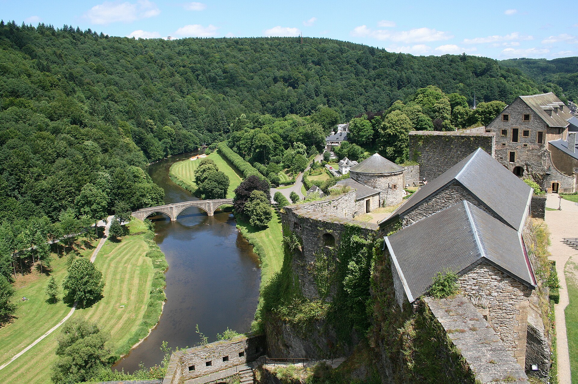 The view from the ramparts of Bouillon castle over the river Semois, an arched stone bridge and wooded hills, with the castle's slate roofs and round tower in the foreground