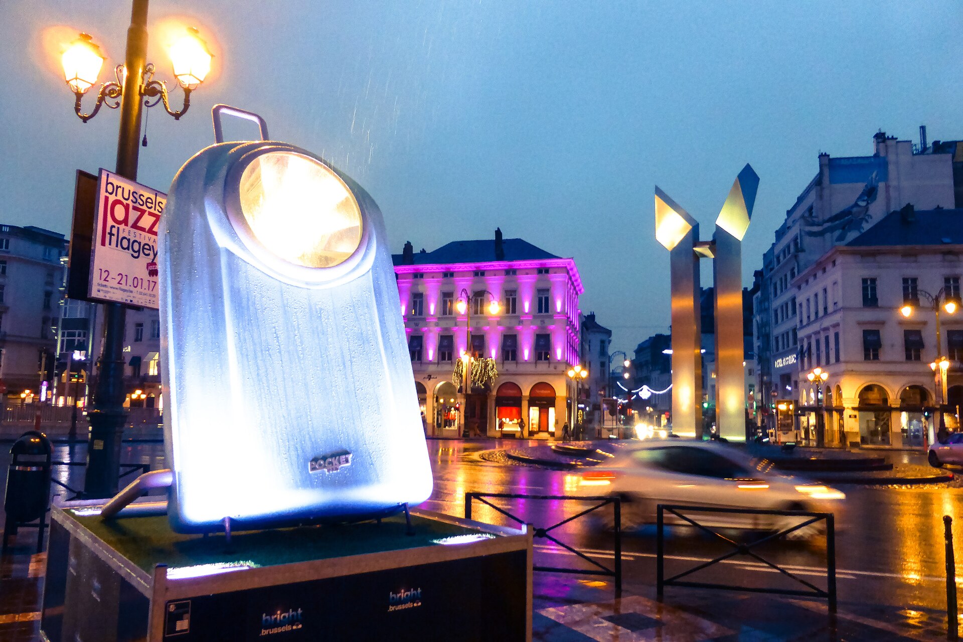 An illuminated light-festival installation on a wet Brussels street at dusk with buildings lit in pink behind