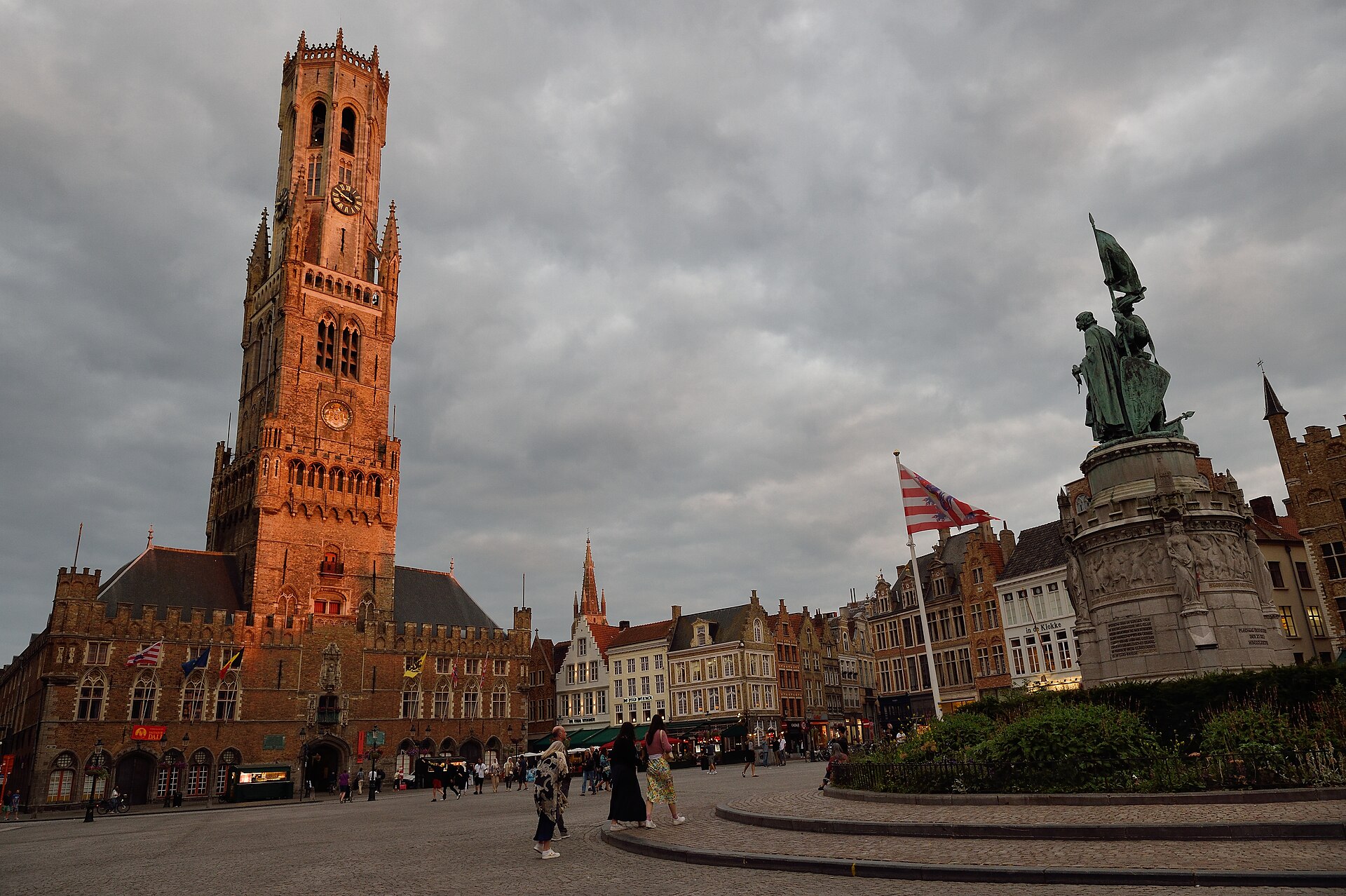The Belfry of Bruges lit by evening light above a near-empty Markt square with the Jan Breydel statue in the foreground