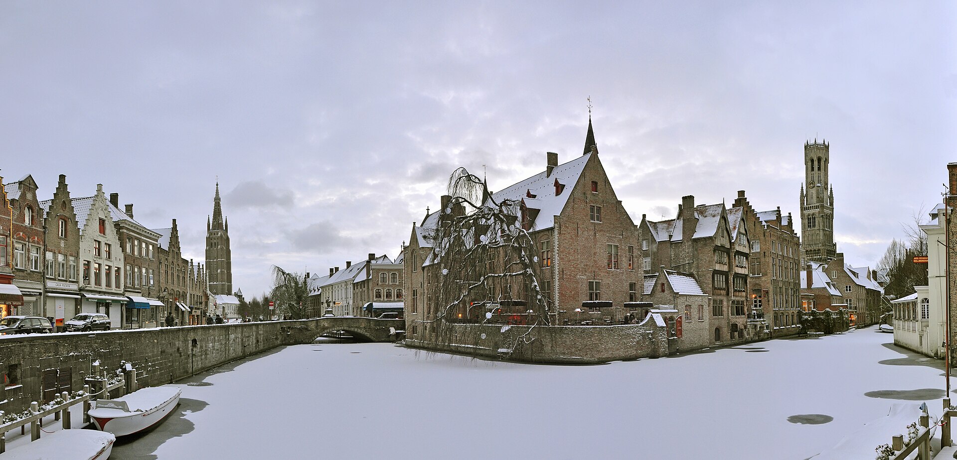A frozen canal at the Rozenhoedkaai in Bruges under snow, with the Belfry and the tower of the Church of Our Lady behind