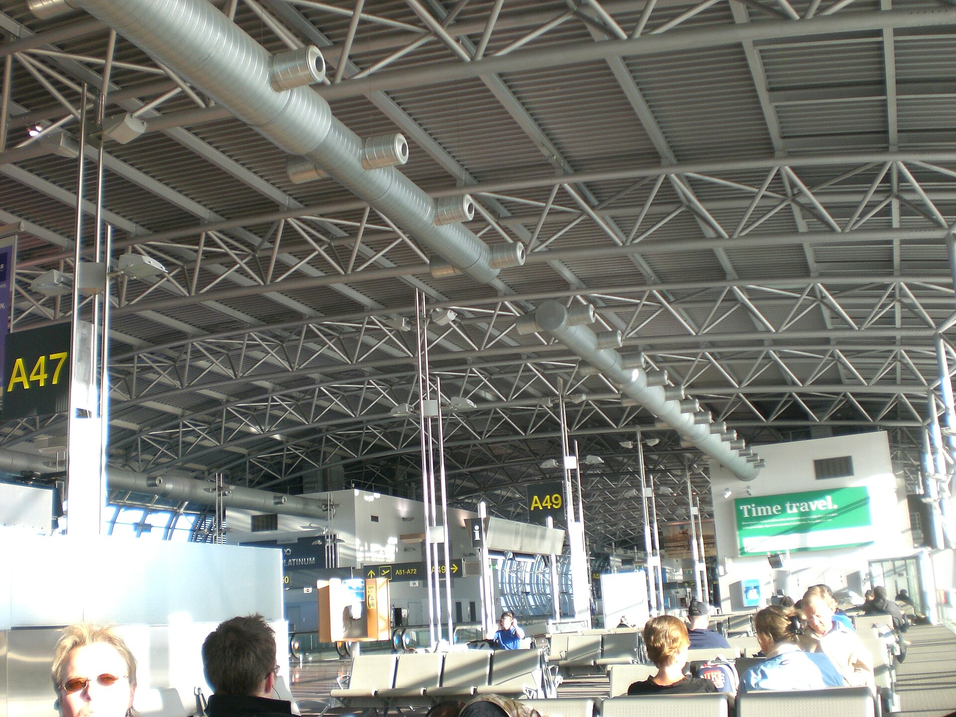 The barrel-vaulted steel roof of Pier A at Brussels Airport with gate signs for A47 and A49 above seated passengers