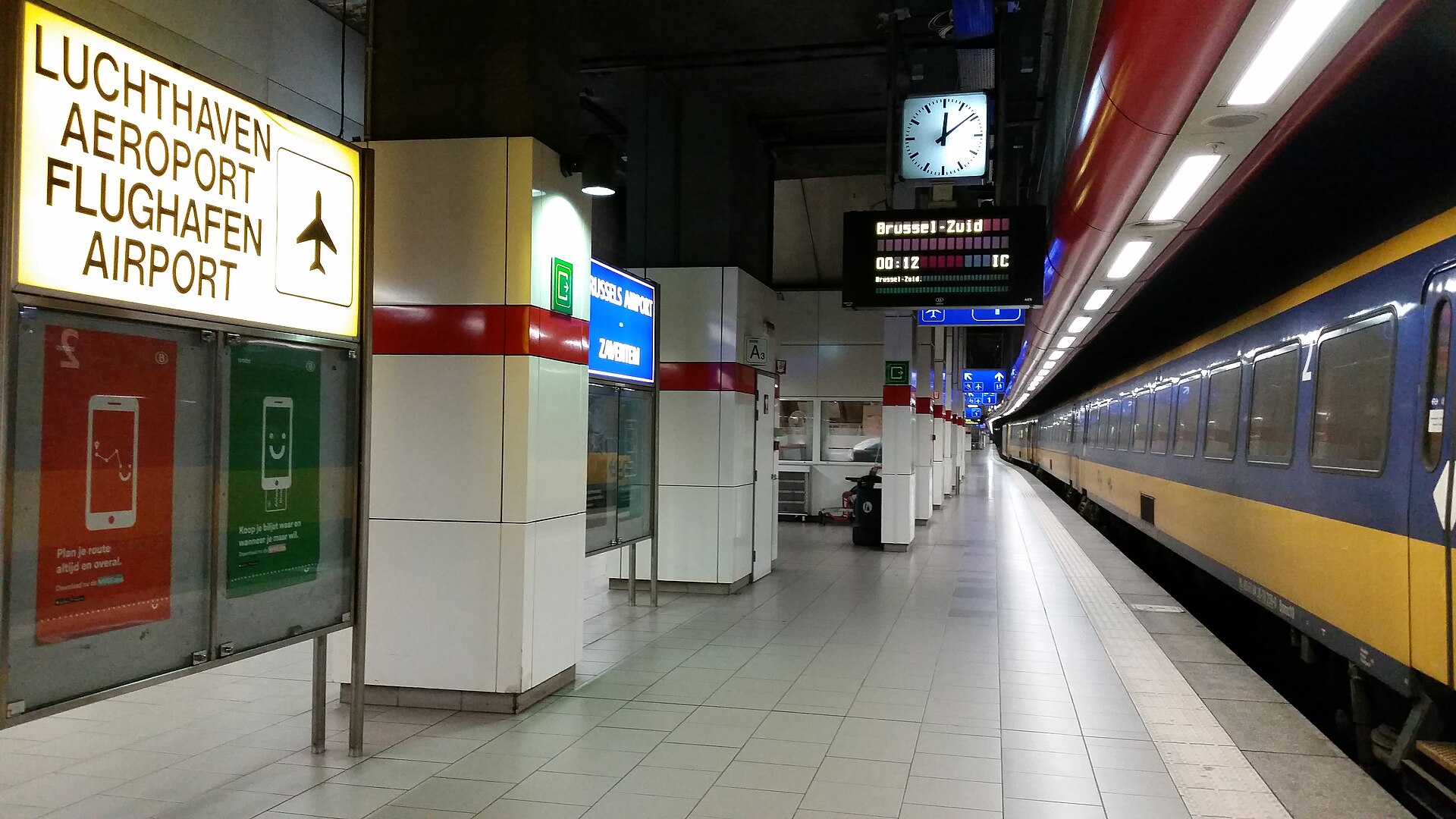 The underground railway platform at Brussels Airport with a quadrilingual airport sign and a departure board showing a train to Brussel-Zuid