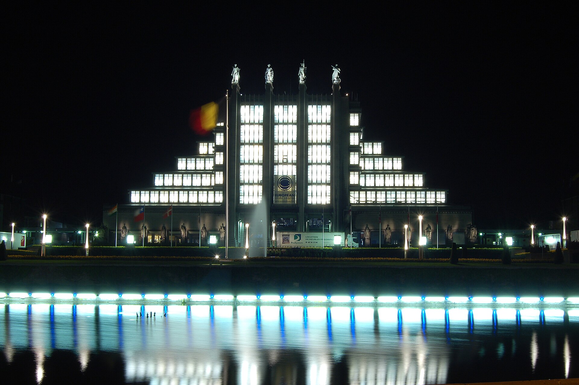 The floodlit stepped facade of the Palais du Centenaire at Brussels Expo reflected in water at night