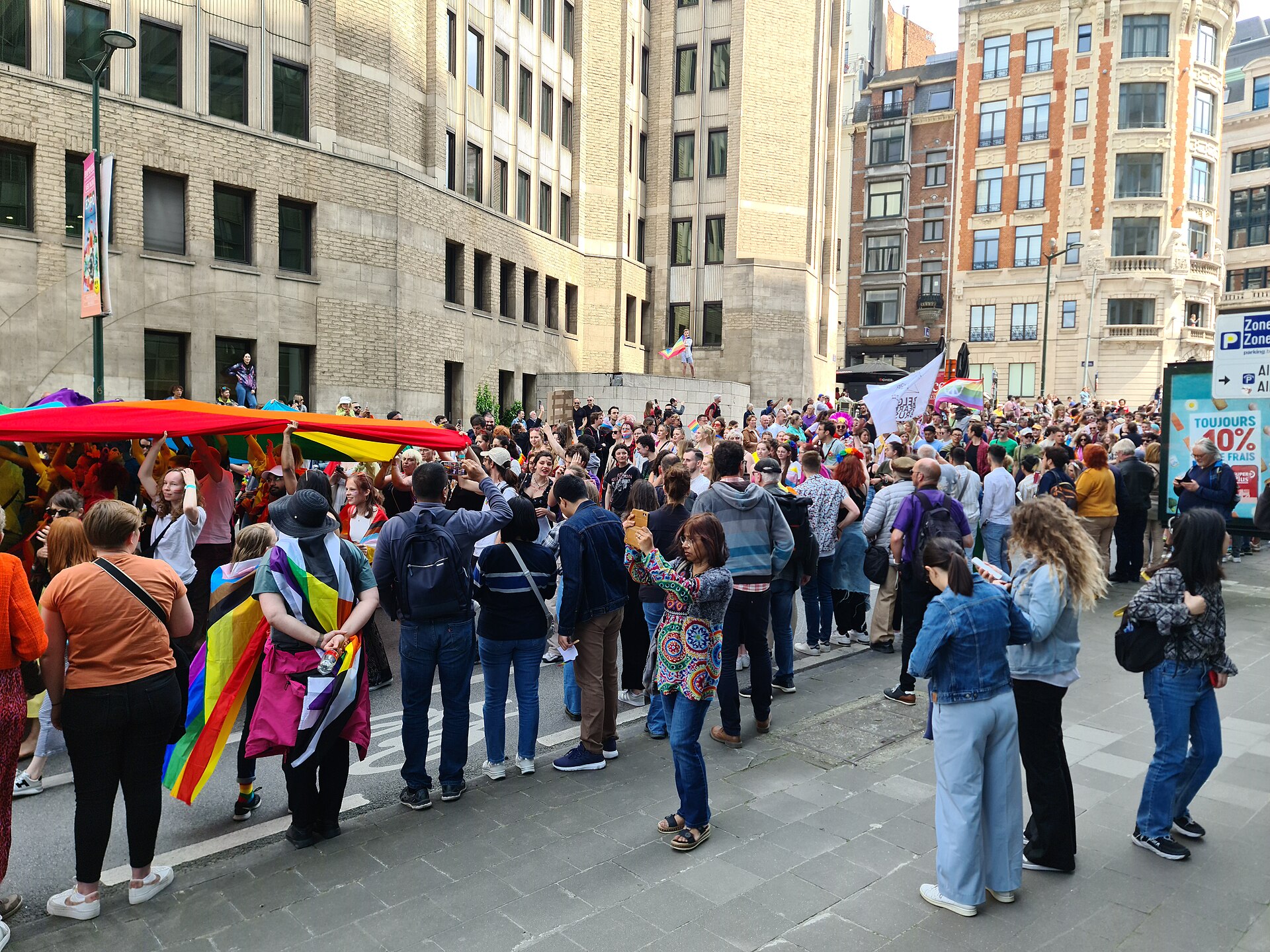 Crowds with rainbow and progress pride flags gathering in a sunny street in central Brussels during the Pride parade, with a long rainbow banner held overhead