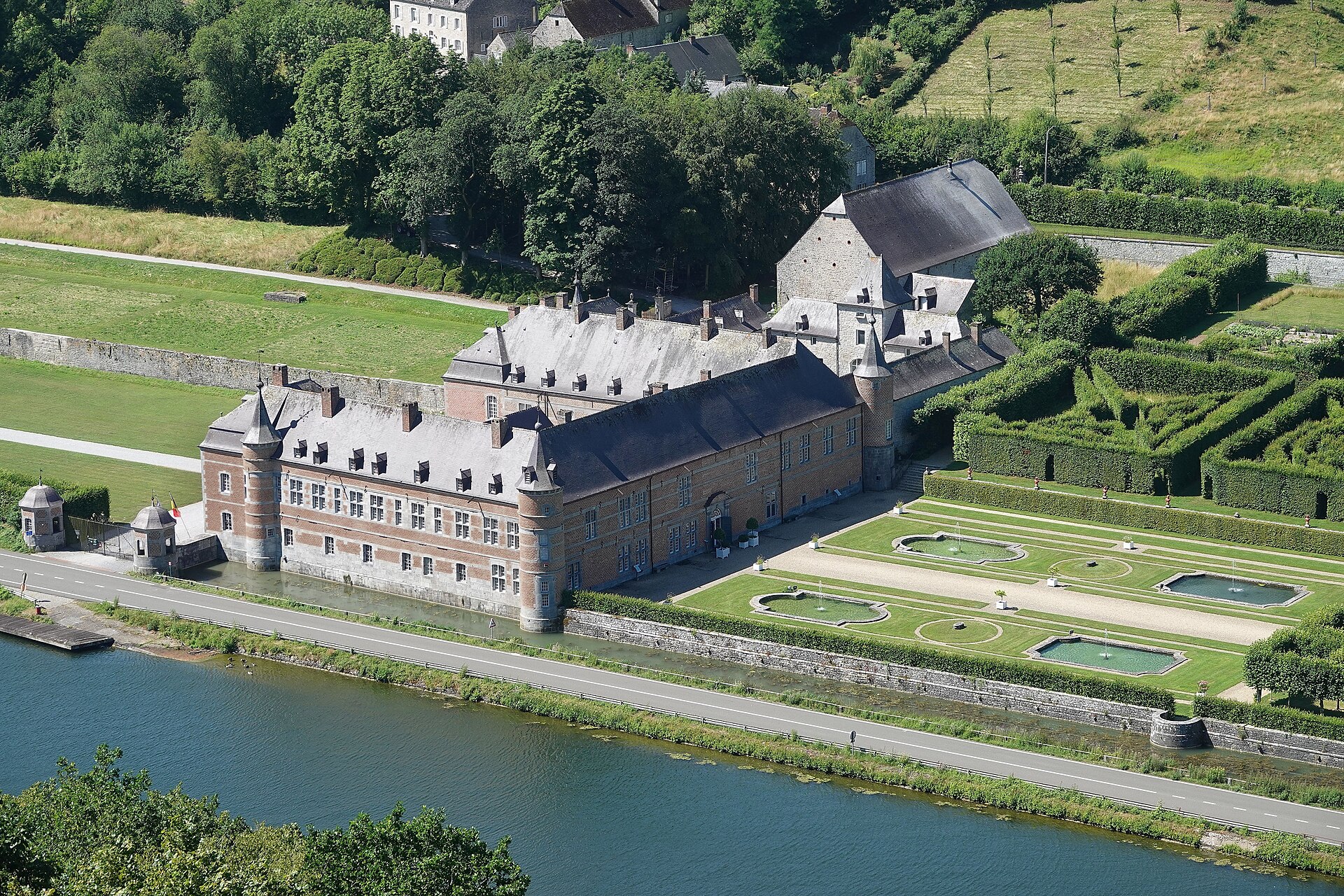 Aerial view of the brick and stone Château de Freÿr on the bank of the Meuse, with its formal gardens, fountain basins and hedge maze beside the river road