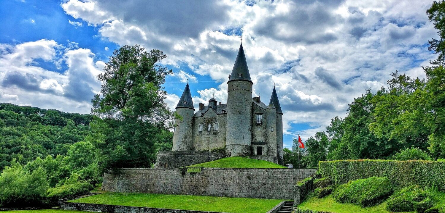 The Château de Vêves with its round stone towers and tall pointed slate roofs on a terraced green mound under a cloudy blue sky