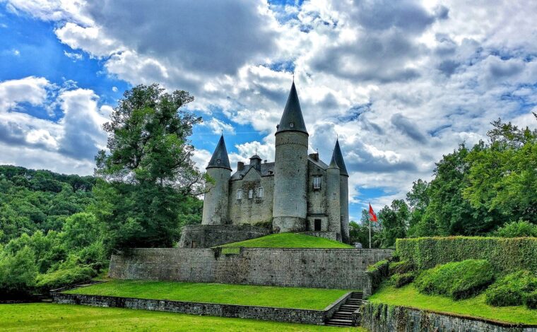 The Château de Vêves with its round stone towers and tall pointed slate roofs on a terraced green mound under a cloudy blue sky