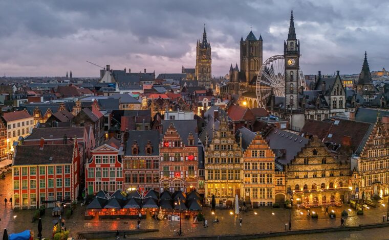 An aerial view of the lit guild houses of the Graslei in Ghent at dusk, with the Belfry, Saint Nicholas' Church, the old post office clock tower and a Ferris wheel behind