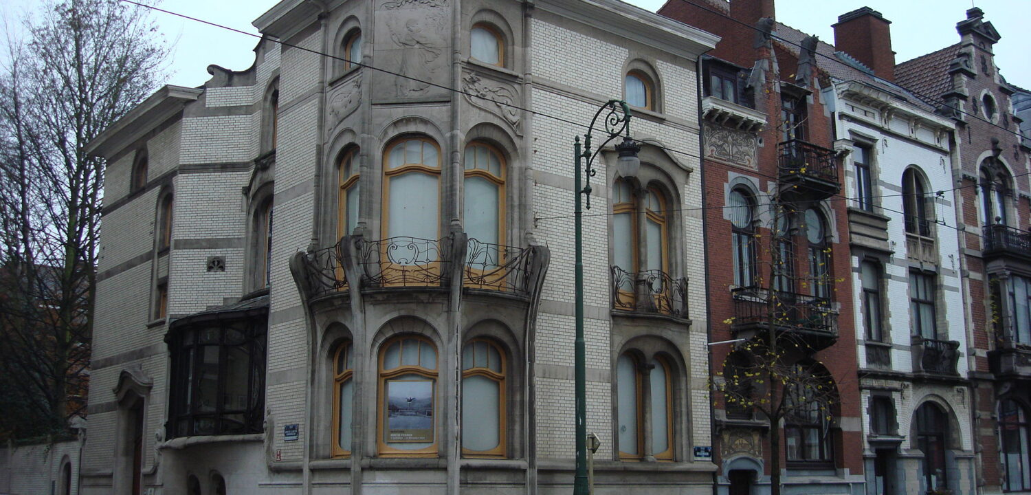The pale Art Nouveau corner facade of the Hôtel Hannon in Saint-Gilles, with curved bay windows and wrought-iron balconies, on a grey day with bare trees