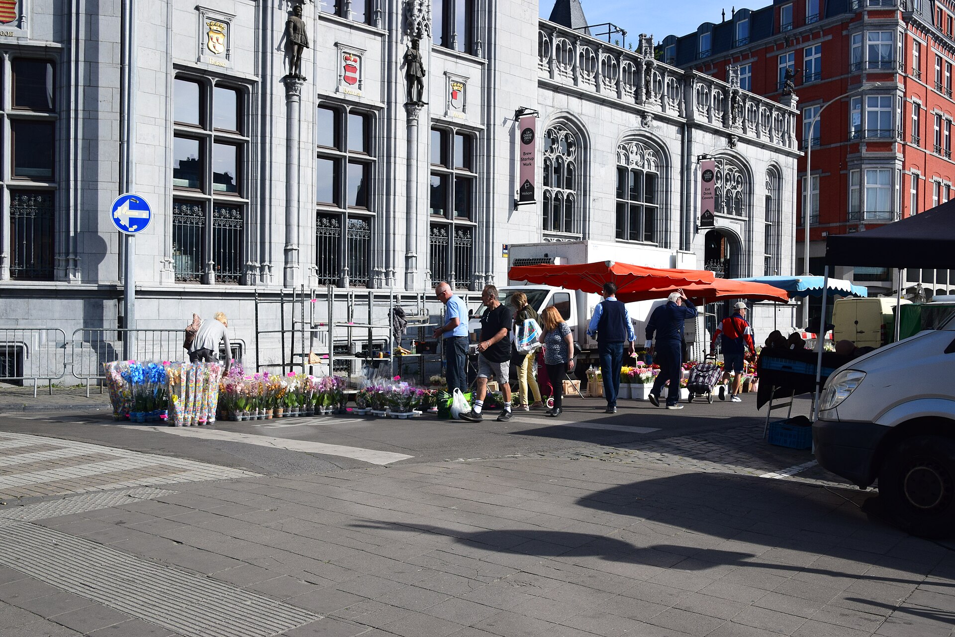 Flower and plant stalls with orange parasols on a Sunday morning at La Batte market, in front of the neo-Gothic stone facade of Liège's former main post office