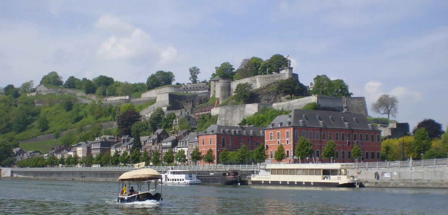 Namur citadel's stone ramparts on the wooded hill above the Meuse, with red-brick buildings on the quay and a small canopied Namourette boat on the river