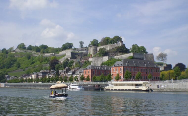 Namur citadel's stone ramparts on the wooded hill above the Meuse, with red-brick buildings on the quay and a small canopied Namourette boat on the river