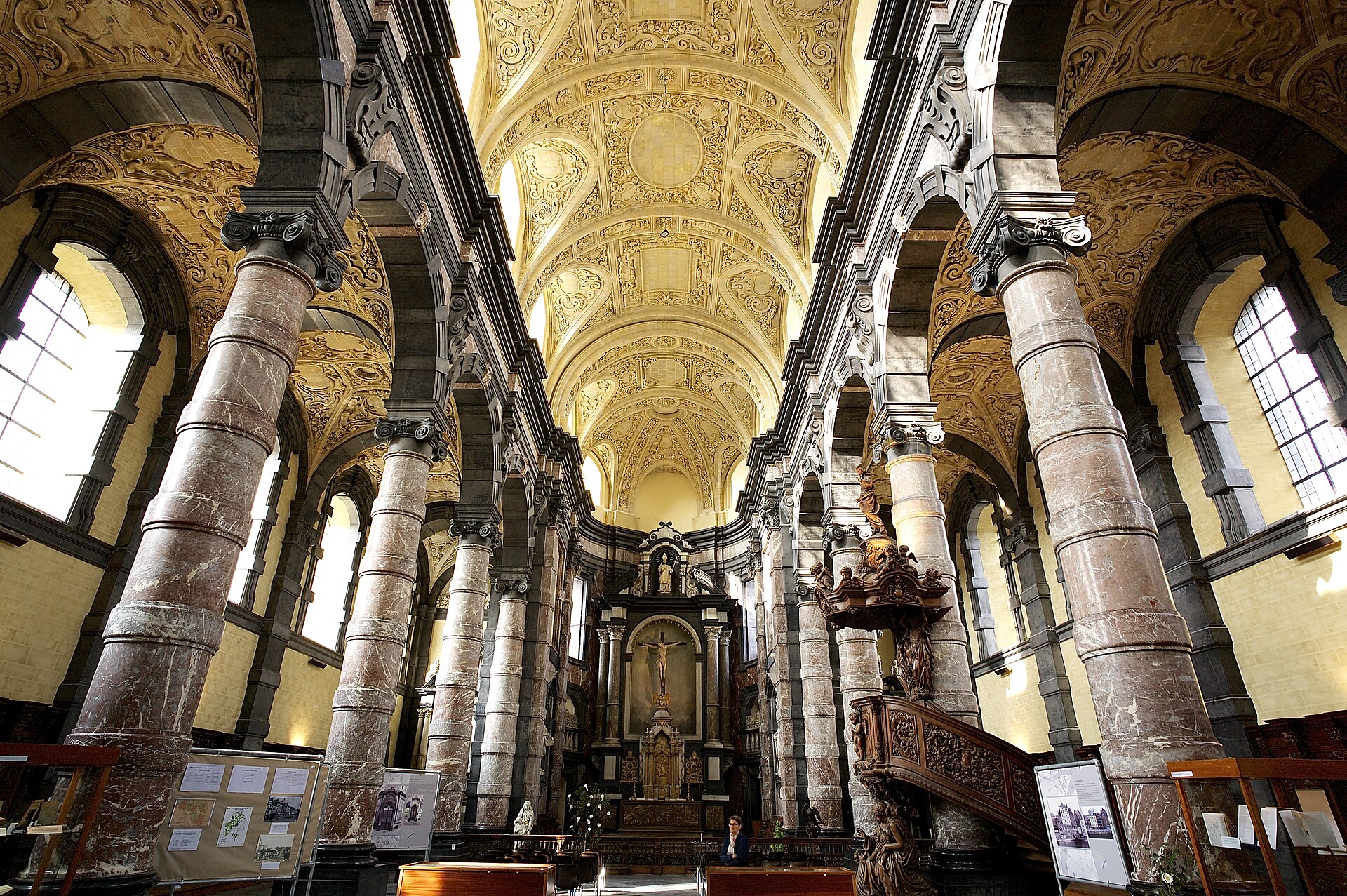 The nave of the Église Saint-Loup in Namur, lined with red marble columns under a pale sculpted stone vault, with a carved wooden pulpit on the right