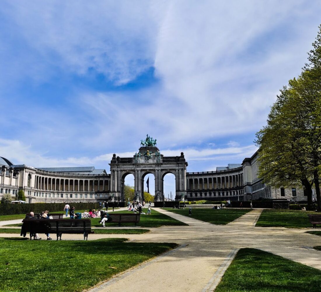 People relaxing on the lawns and benches of the Parc du Cinquantenaire on a bright spring day, with the triumphal arcade and colonnades behind