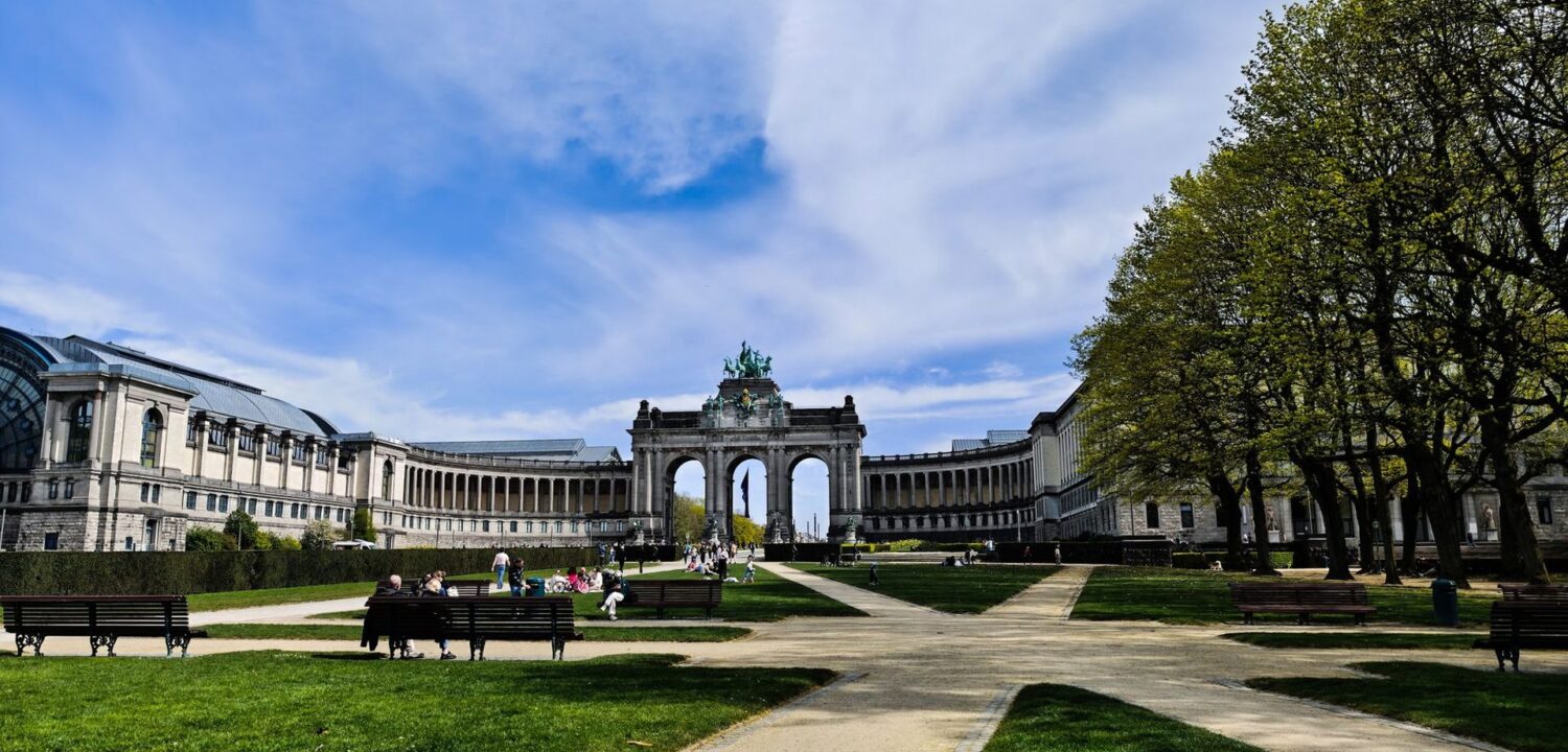 People relaxing on the lawns and benches of the Parc du Cinquantenaire on a bright spring day, with the triumphal arcade and colonnades behind
