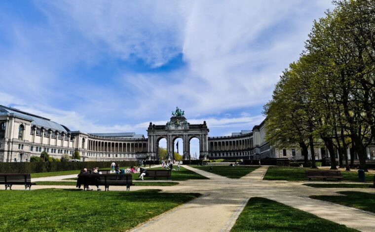 People relaxing on the lawns and benches of the Parc du Cinquantenaire on a bright spring day, with the triumphal arcade and colonnades behind