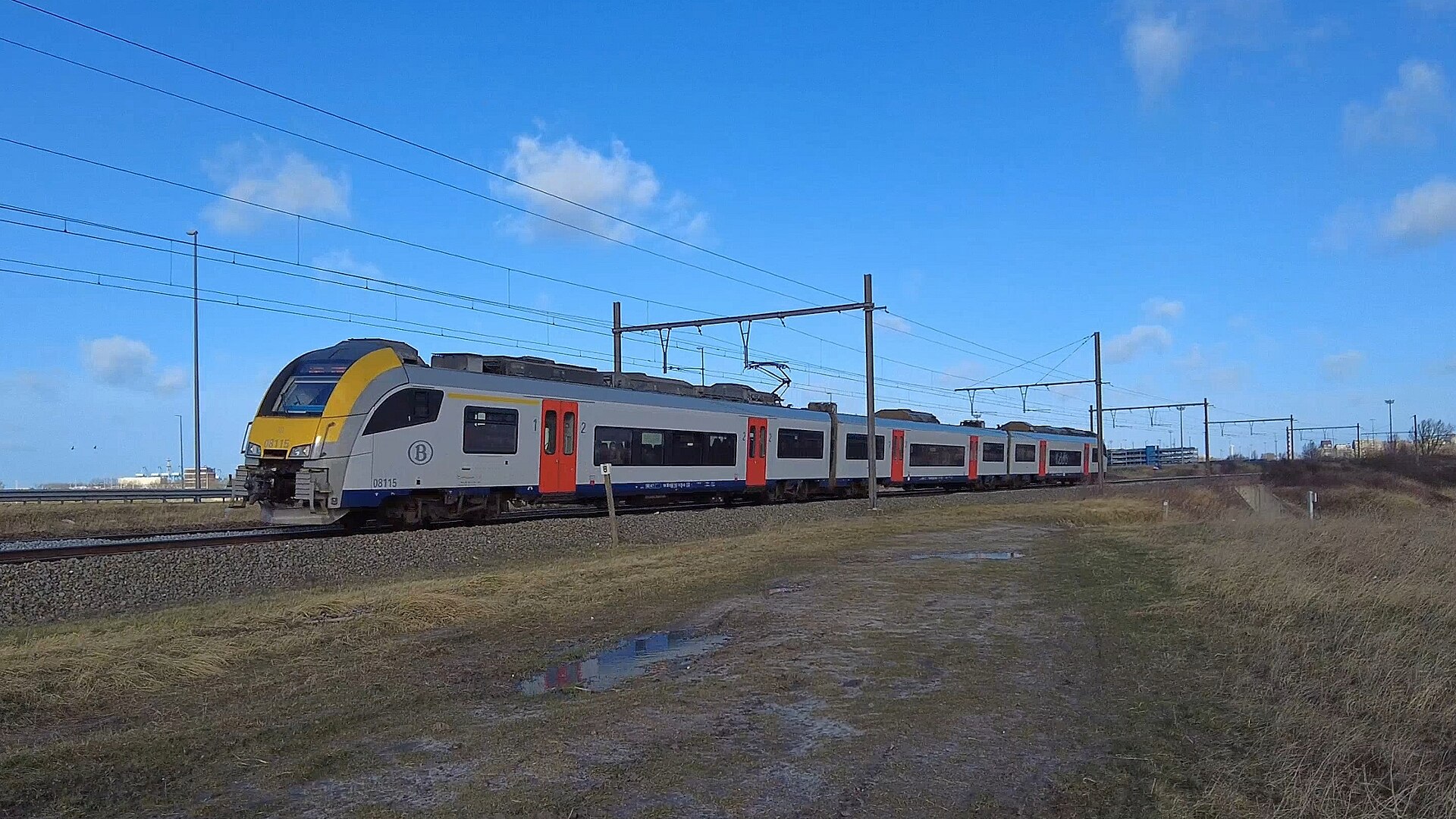 A silver, yellow and blue SNCB Desiro electric train with red doors running under overhead wires across flat open country near the coast