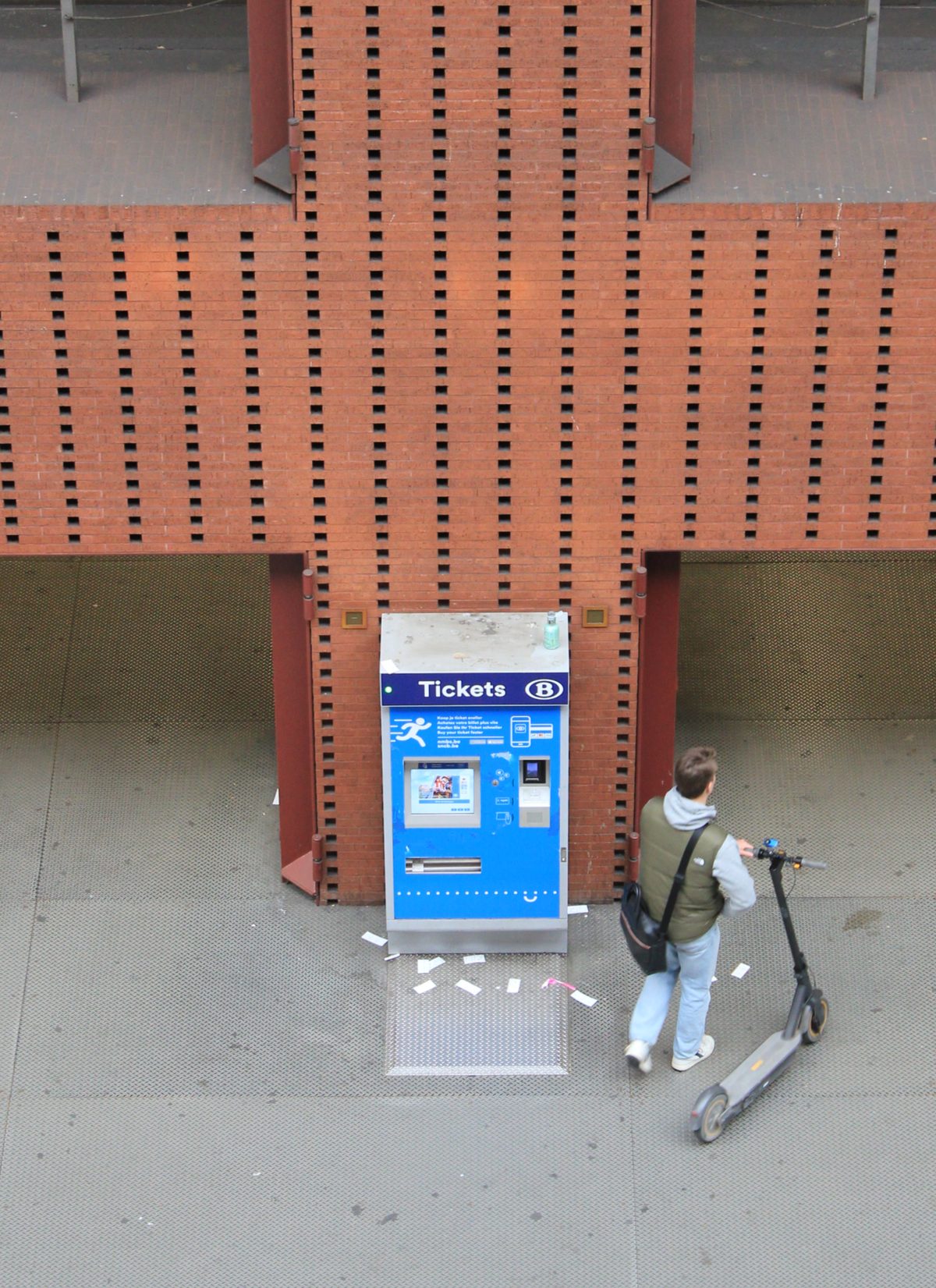 A blue SNCB ticket machine with a card reader and touchscreen against a red-brick pillar, seen from above with a commuter and an e-scooter beside it