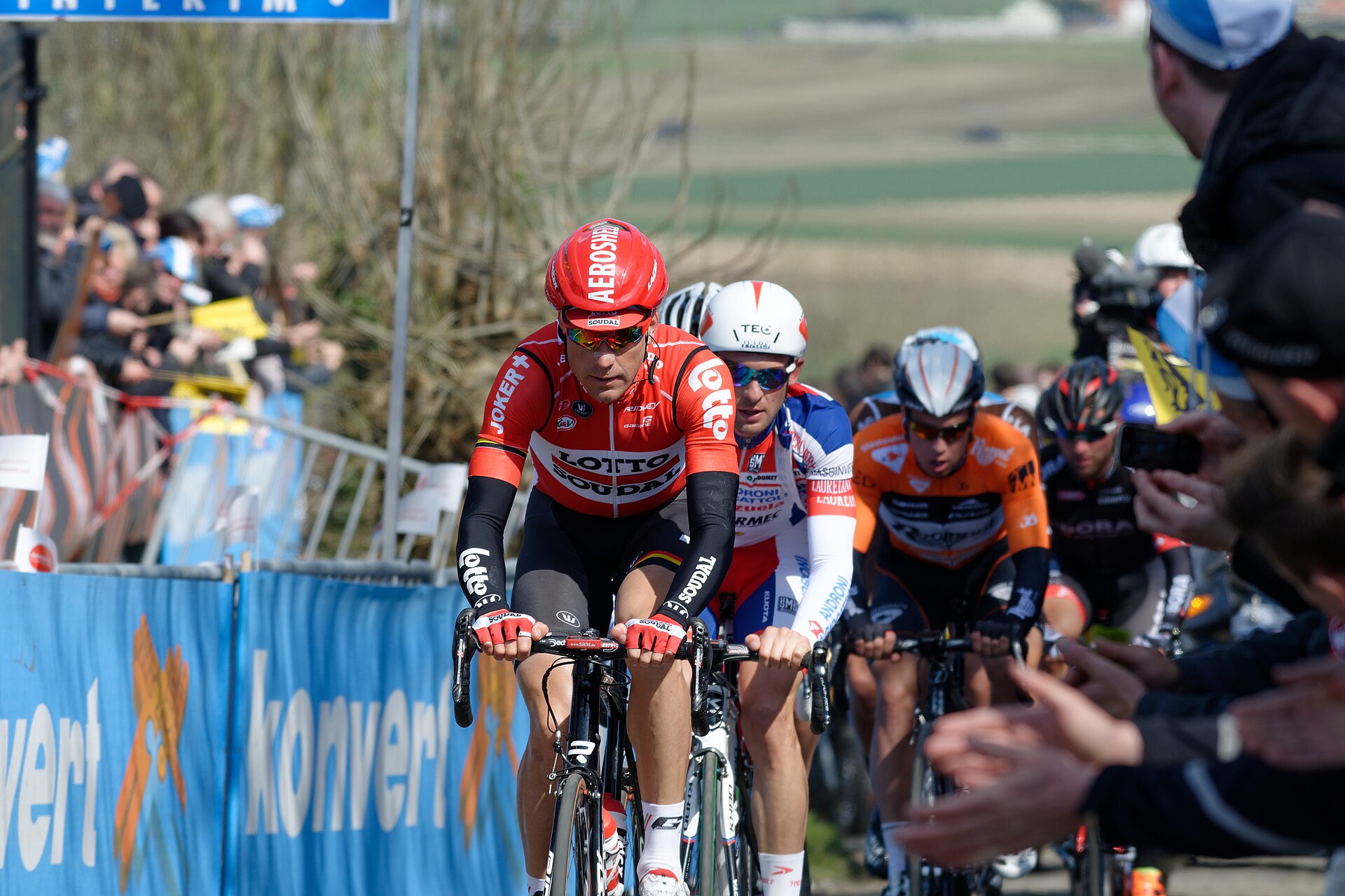 Professional cyclists climbing the Oude Kwaremont during the Tour of Flanders, with fans leaning over the barriers and fields in the background