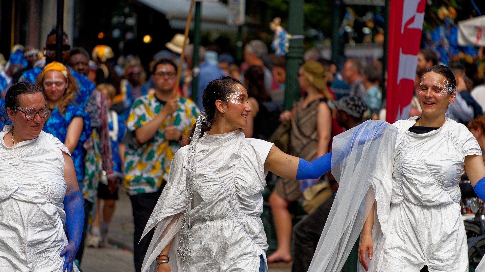 Smiling performers in white paper costumes with blue-painted arms and glittering faces dancing through a crowded street in the Zinneke Parade