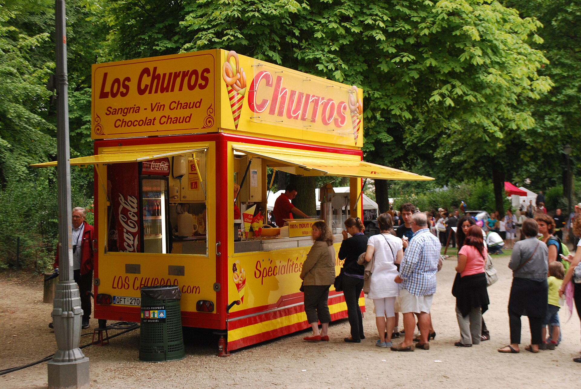 Adults and children queue at a yellow churros stall beneath leafy trees in Brussels Park during Belgian National Day celebrations.