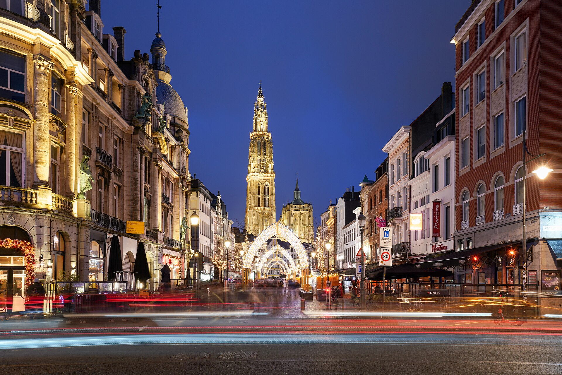 Antwerp’s illuminated cathedral spire rises beyond Suikerrui, where festive light arches and passing traffic brighten the evening street.