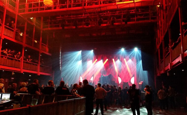 Audience members gather beneath balconies and red stage lights inside the Ancienne Belgique concert hall in Brussels.