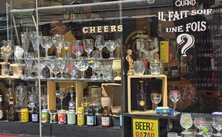 Beerz Bazar in Brussels displays Belgian beer bottles, cans and branded glasses, with densely stocked shelves visible behind the window.
