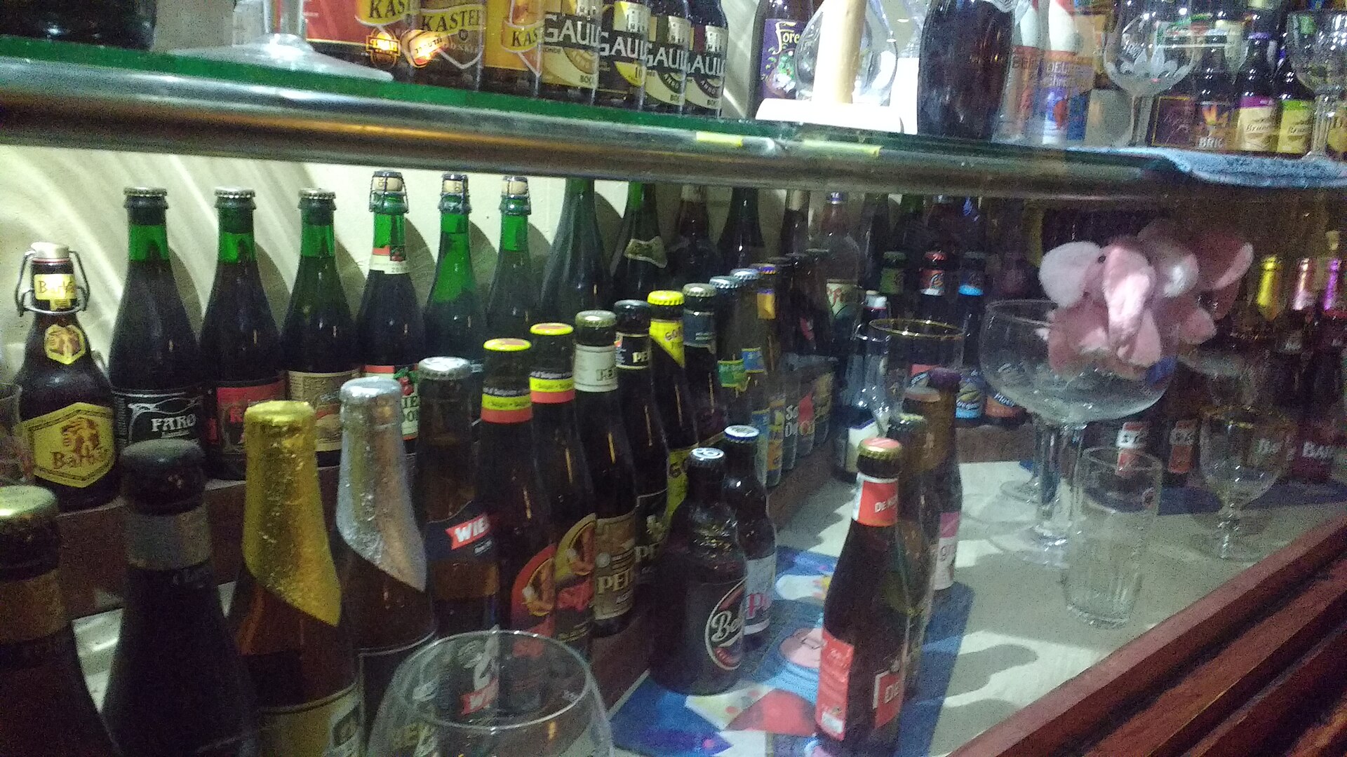 Belgian beer bottles and a glass stand before refrigerated shelves at Delirium Cafe in Brussels, beside a pink elephant mascot.