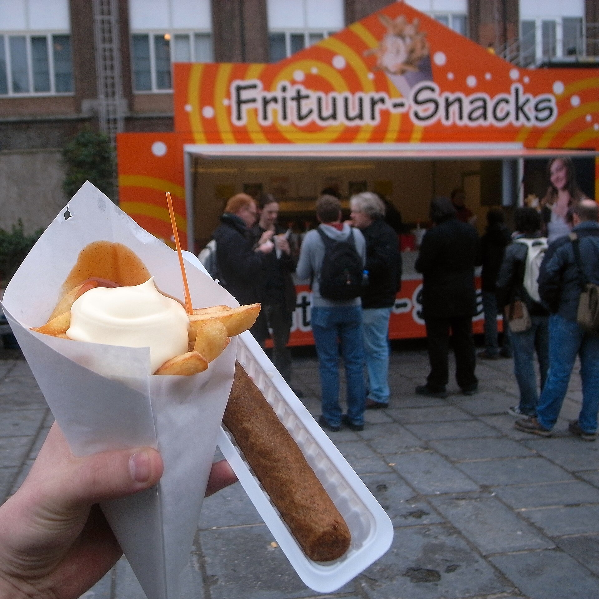 Belgian frites topped with mayonnaise fill a paper cone beside a frikandel, with customers waiting at a frituur behind.