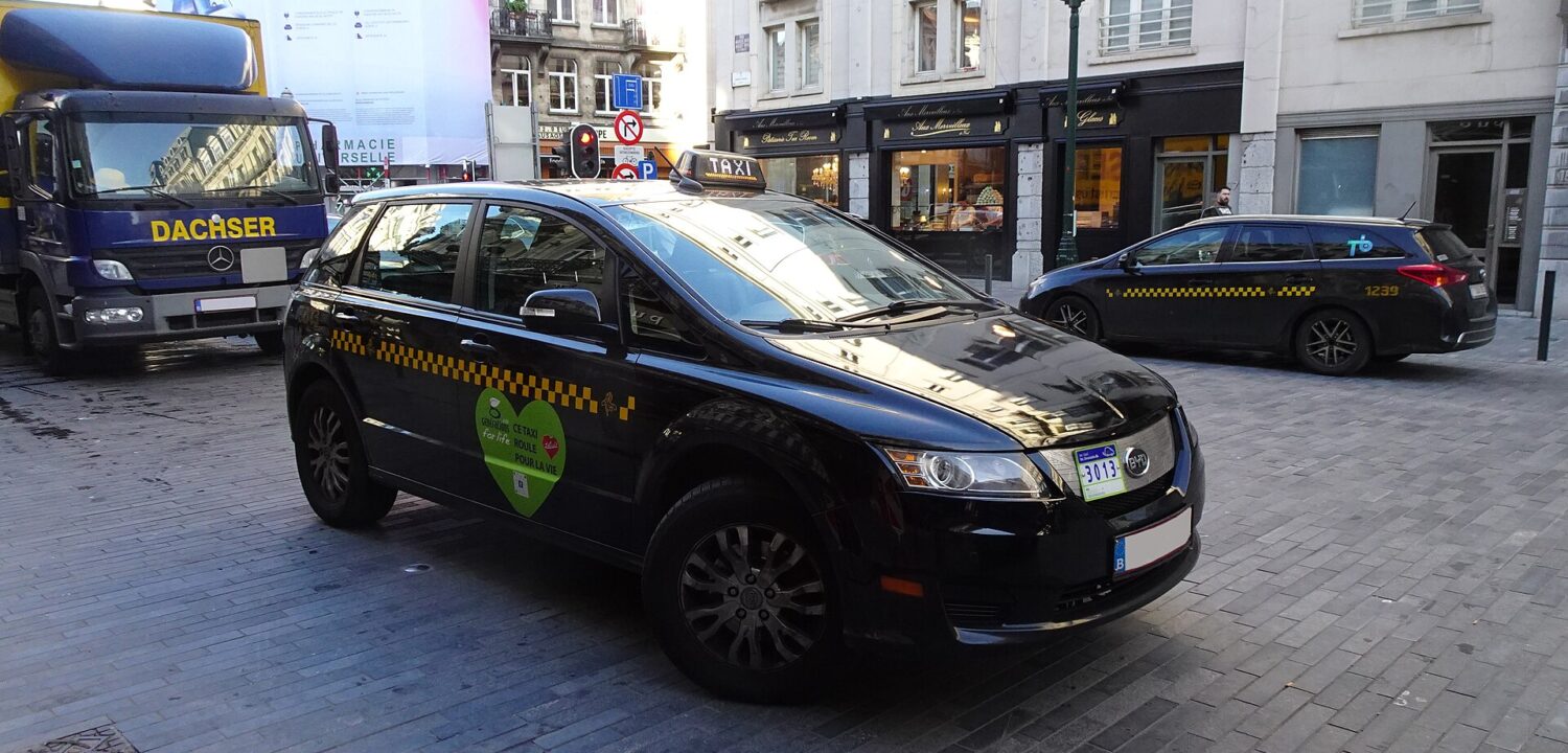 A black electric taxi with yellow checker markings and a roof sign travels along a cobbled Brussels street.