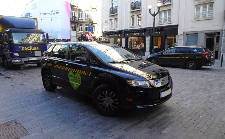 A black electric taxi with yellow checker markings and a roof sign travels along a cobbled Brussels street.
