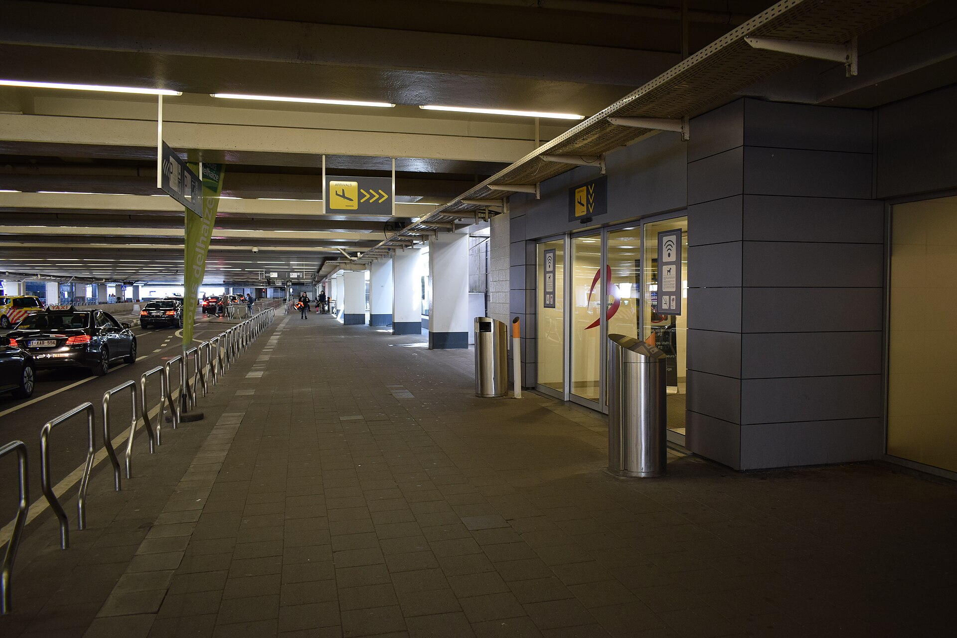 Black taxis queue beneath the covered roadway outside Brussels Airport arrivals, beside metal railings and yellow signs marking the terminal entrance.