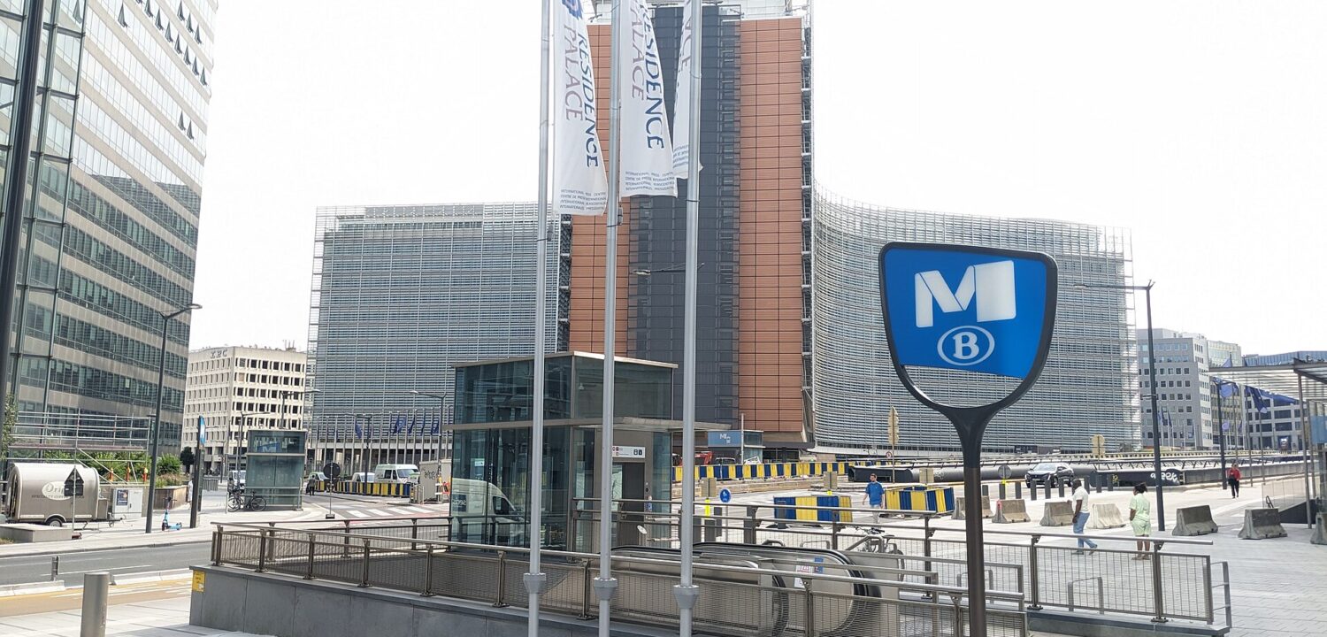 A blue metro M sign marks the Schuman station entrance in Brussels, with escalators descending beside the Berlaymont building in the background.