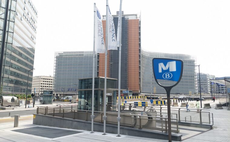 A blue metro M sign marks the Schuman station entrance in Brussels, with escalators descending beside the Berlaymont building in the background.