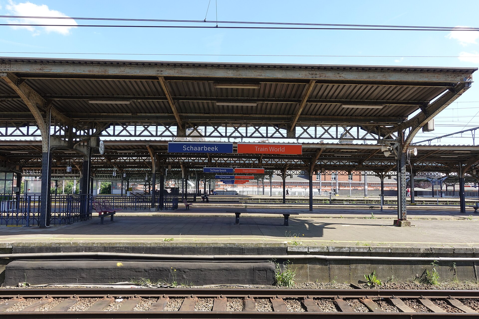 Blue Schaarbeek and orange Train World signs hang beneath iron canopies above the tracks and platforms at Schaerbeek station in Brussels.