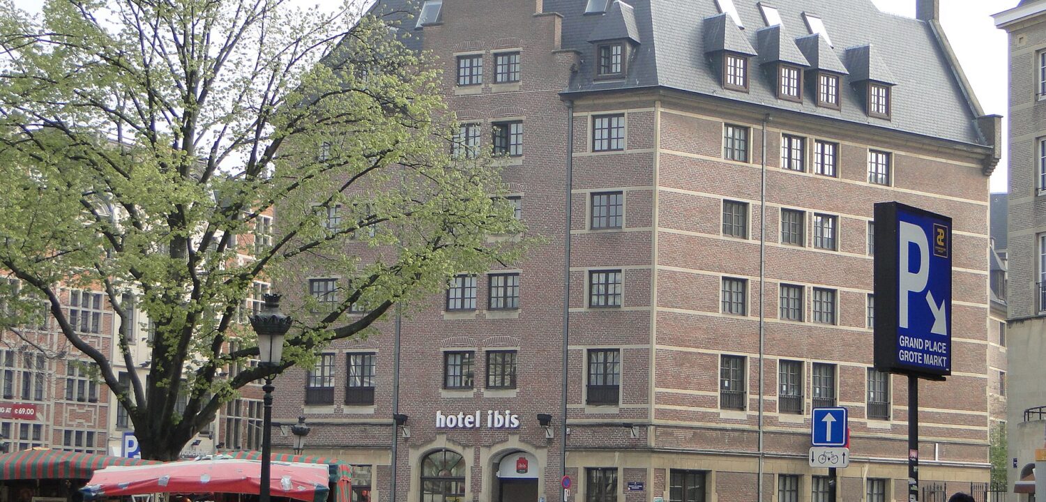 The brick Ibis hotel near Brussels Grand-Place overlooks a cobbled square with market stalls, pedestrians and leafy trees.