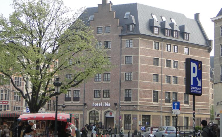 The brick Ibis hotel near Brussels Grand-Place overlooks a cobbled square with market stalls, pedestrians and leafy trees.