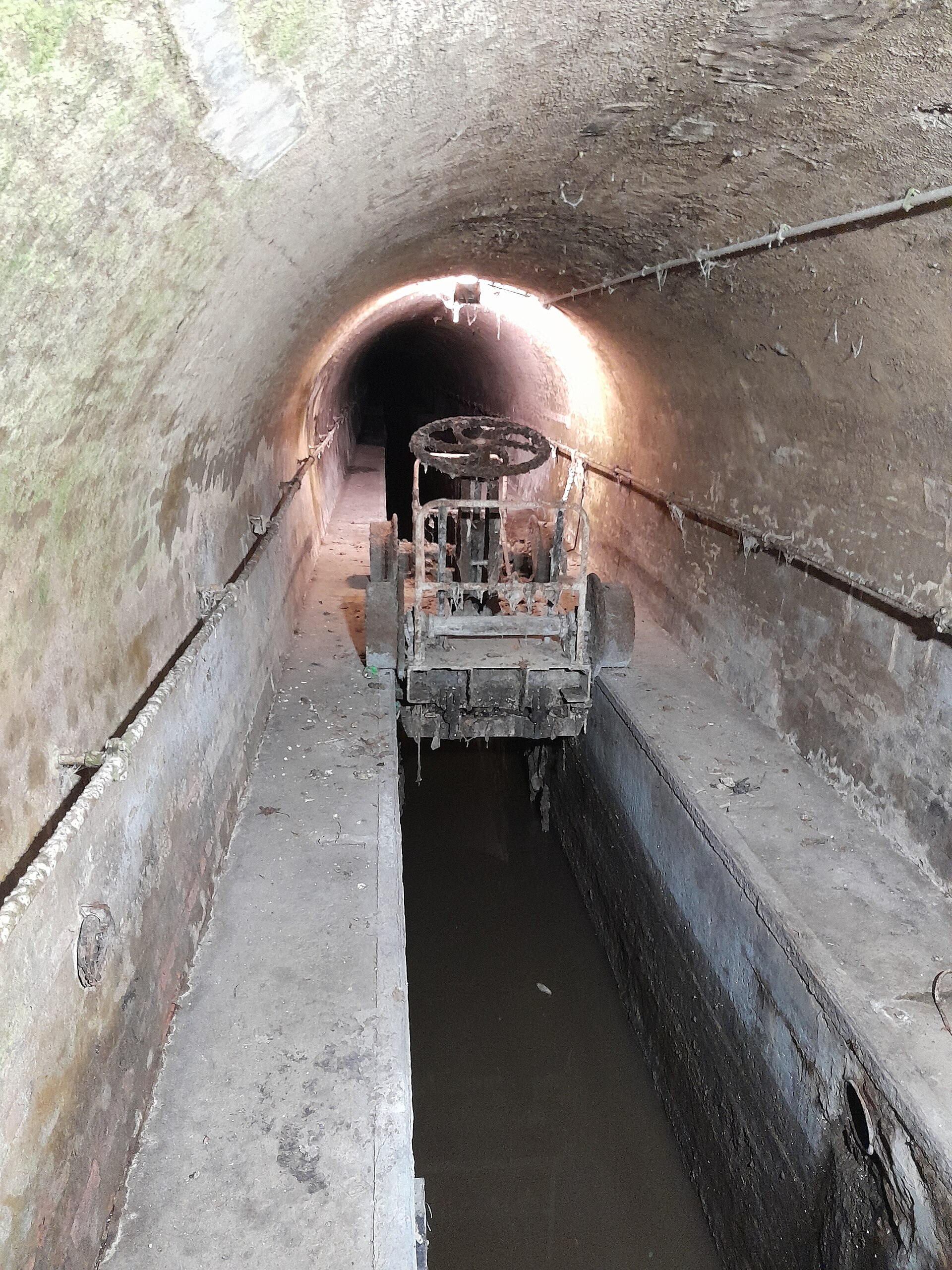 A brick vaulted tunnel at the Brussels Sewer Museum contains a narrow water channel and a wheeled maintenance mechanism.