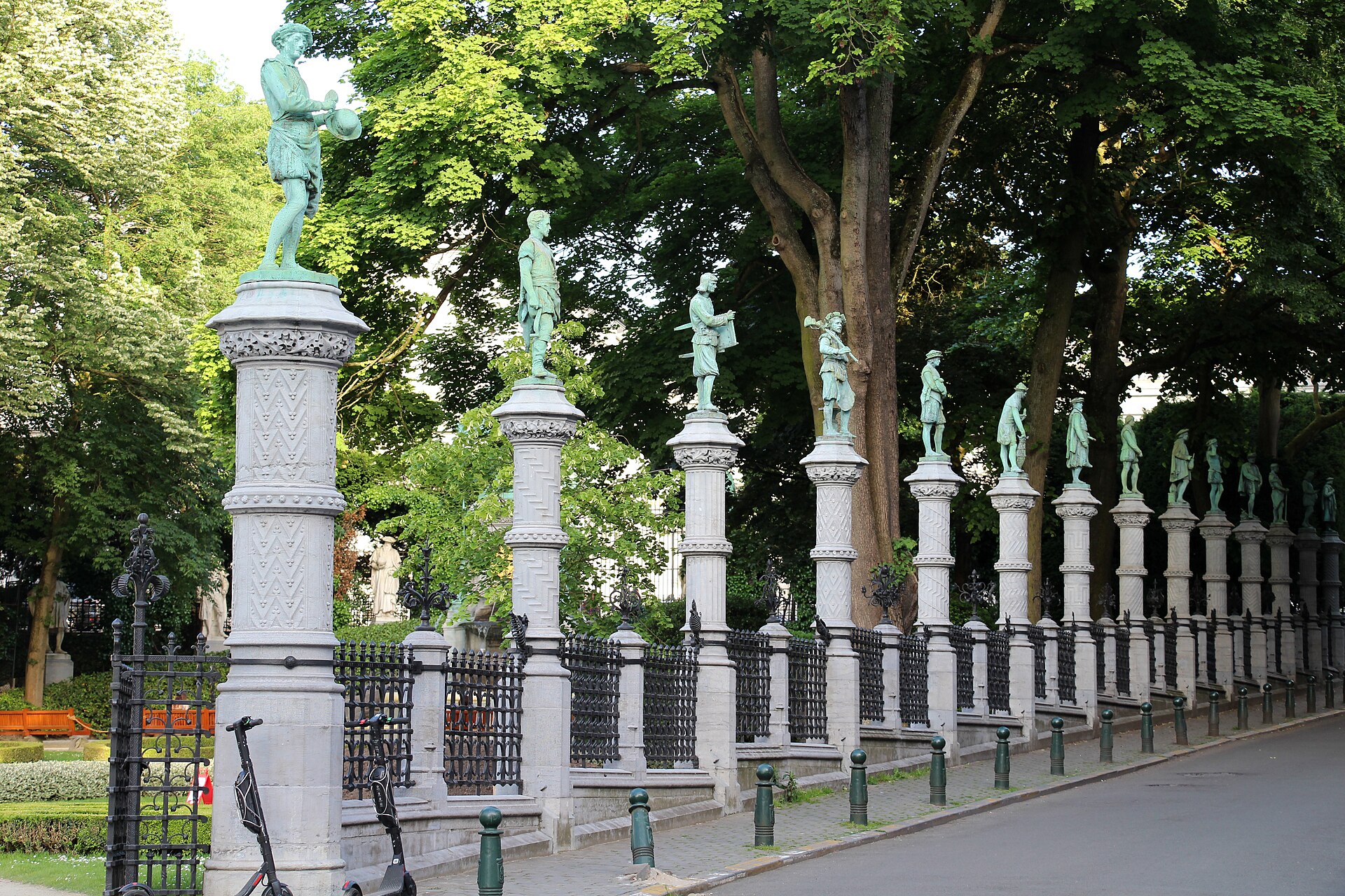 Bronze figures stand on carved stone pillars along the leafy perimeter of the Petit Sablon garden in Brussels.