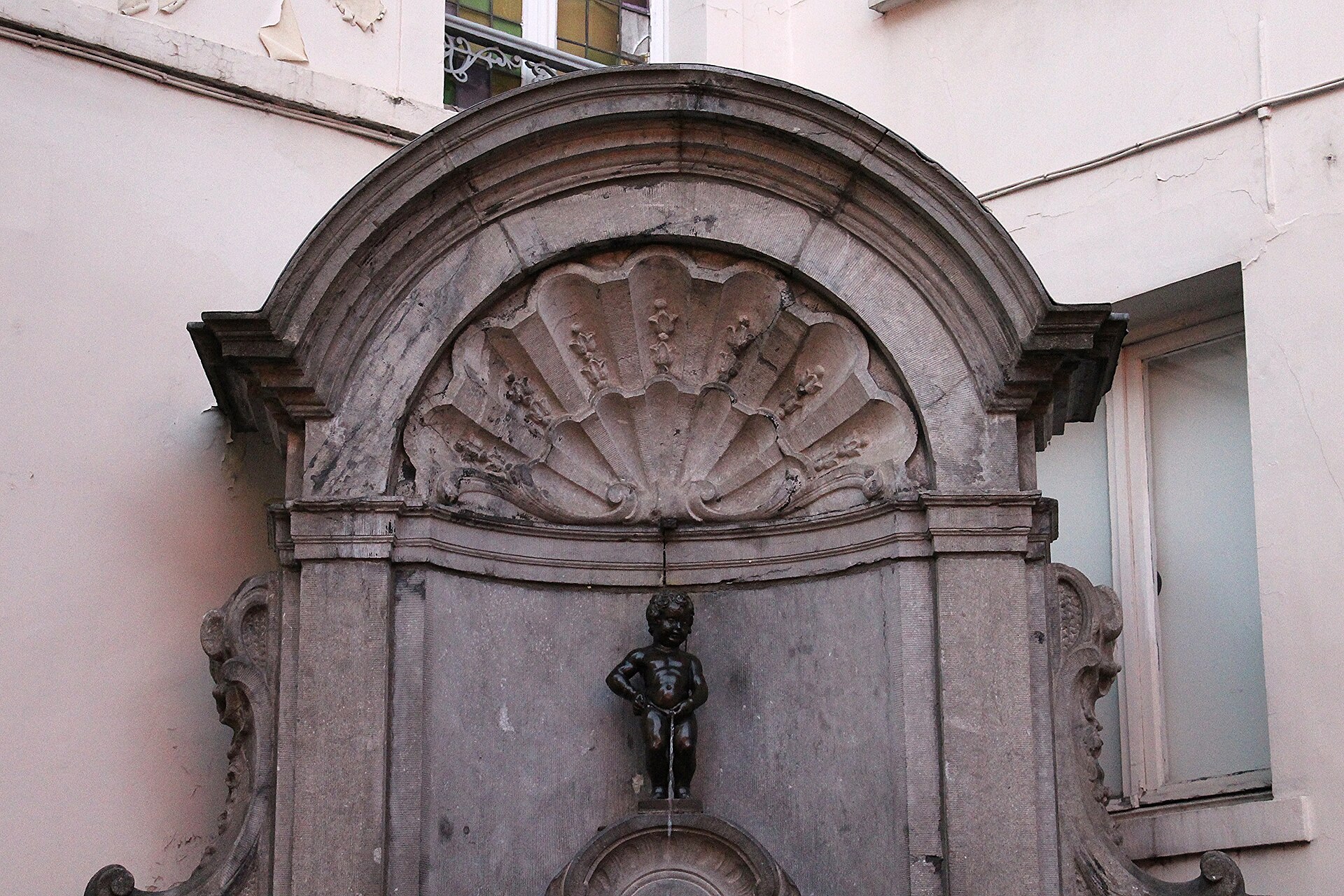 The bronze Manneken Pis fountain stands beneath a carved stone shell in its Brussels street corner niche.