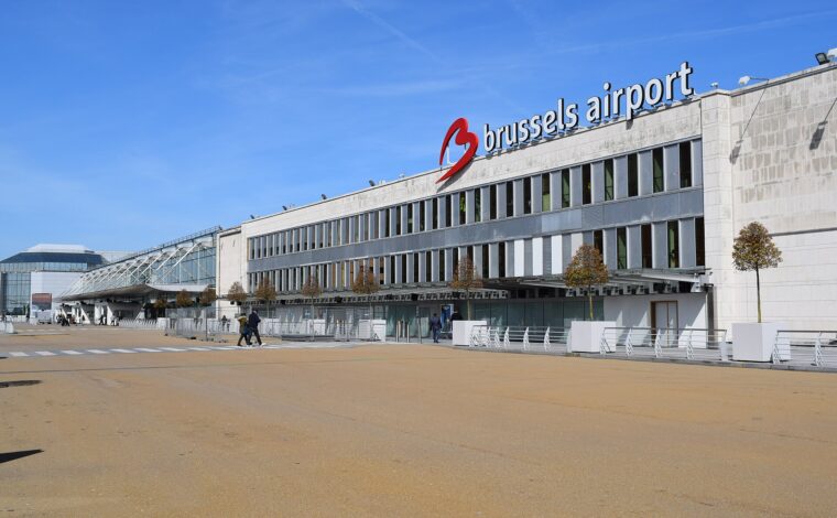 The Brussels Airport name and red logo rise above the terminal facade as travellers cross the sunny forecourt.