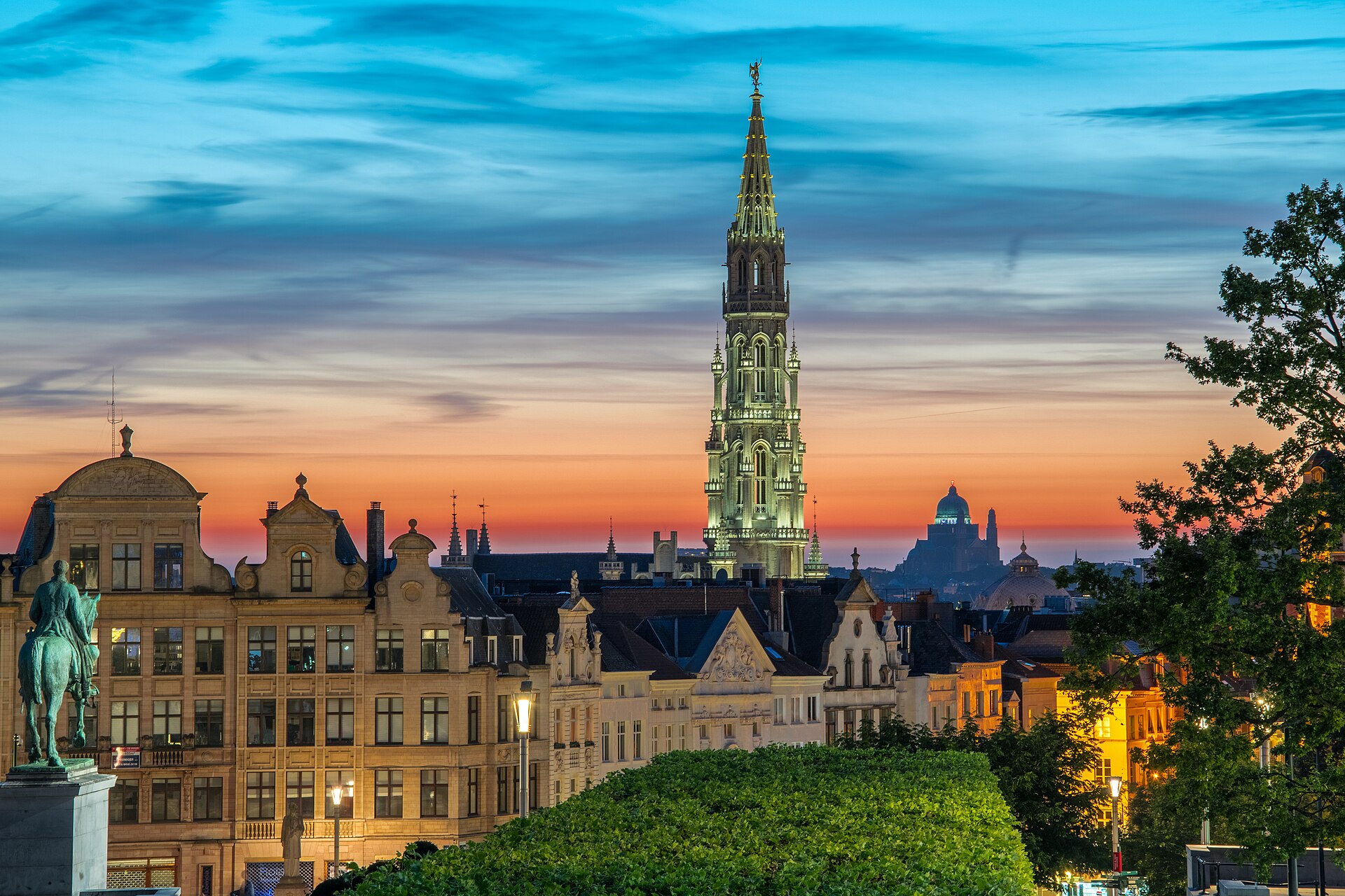Brussels Town Hall glows above historic rooftops at sunset, with the Koekelberg basilica visible beyond the Mont des Arts trees.