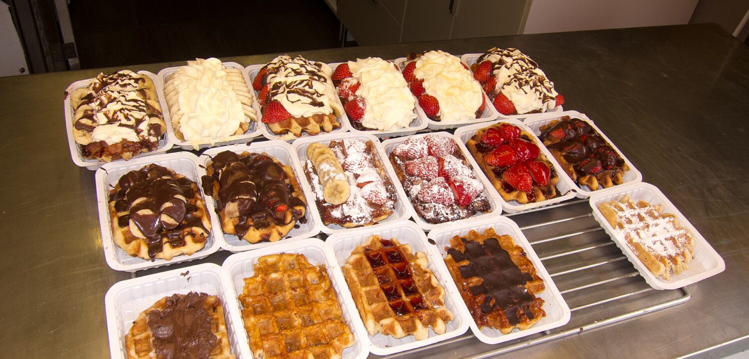 A Brussels waffle counter displays rows of waffles topped with chocolate, strawberries, bananas and whipped cream in individual white trays.