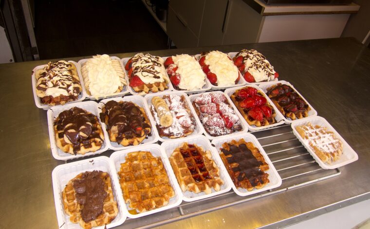 A Brussels waffle counter displays rows of waffles topped with chocolate, strawberries, bananas and whipped cream in individual white trays.