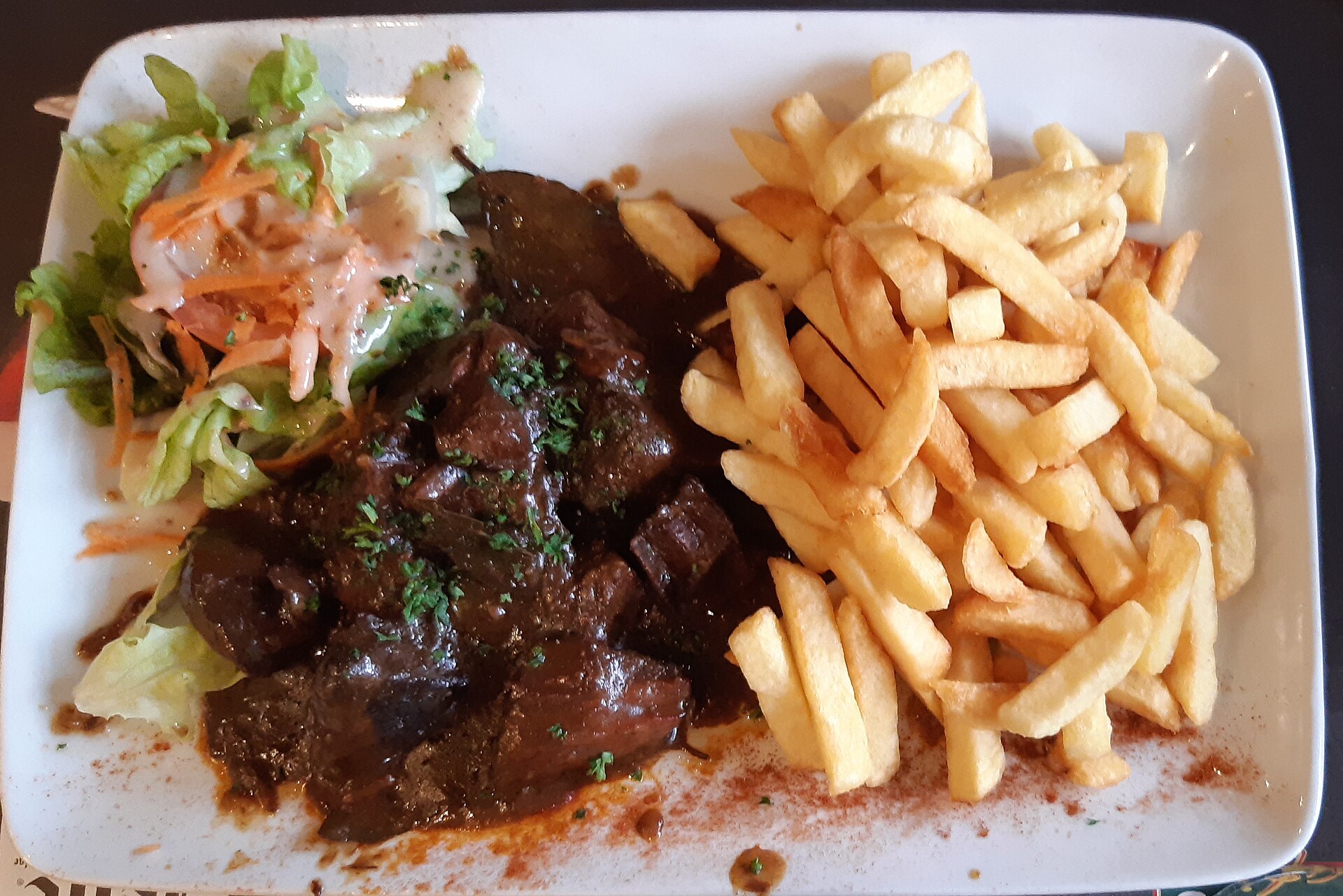 Carbonnade flamande in dark gravy is served with golden fries and a small dressed salad on a rectangular white plate.
