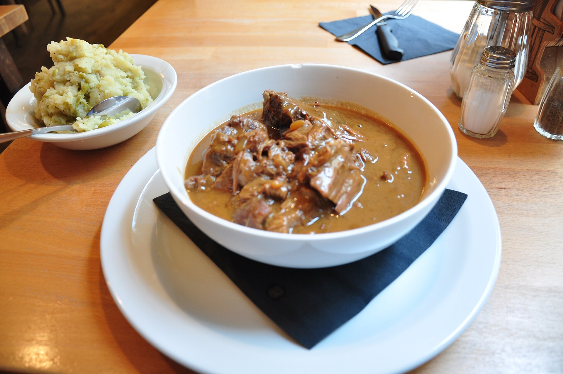 Carbonnade flamande in a white bowl beside mashed potatoes, with cutlery and condiments on a wooden restaurant table.