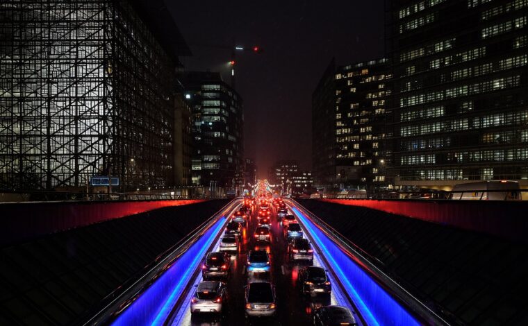 Cars queue at the Belliard tunnel exit in Brussels at night, beneath illuminated European quarter buildings and blue roadside lights.